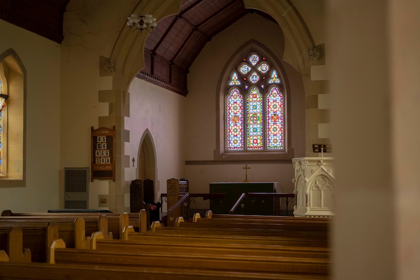 Pews and a stained-glass window are seen inside a stone church.