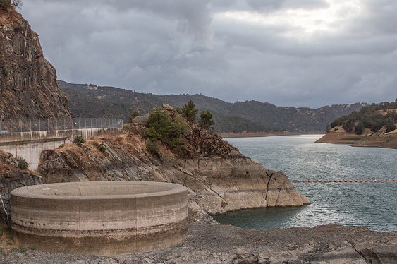 A giant concrete spillway with a dam wall in the background, with a lake with not much water in it and mountains and clouds