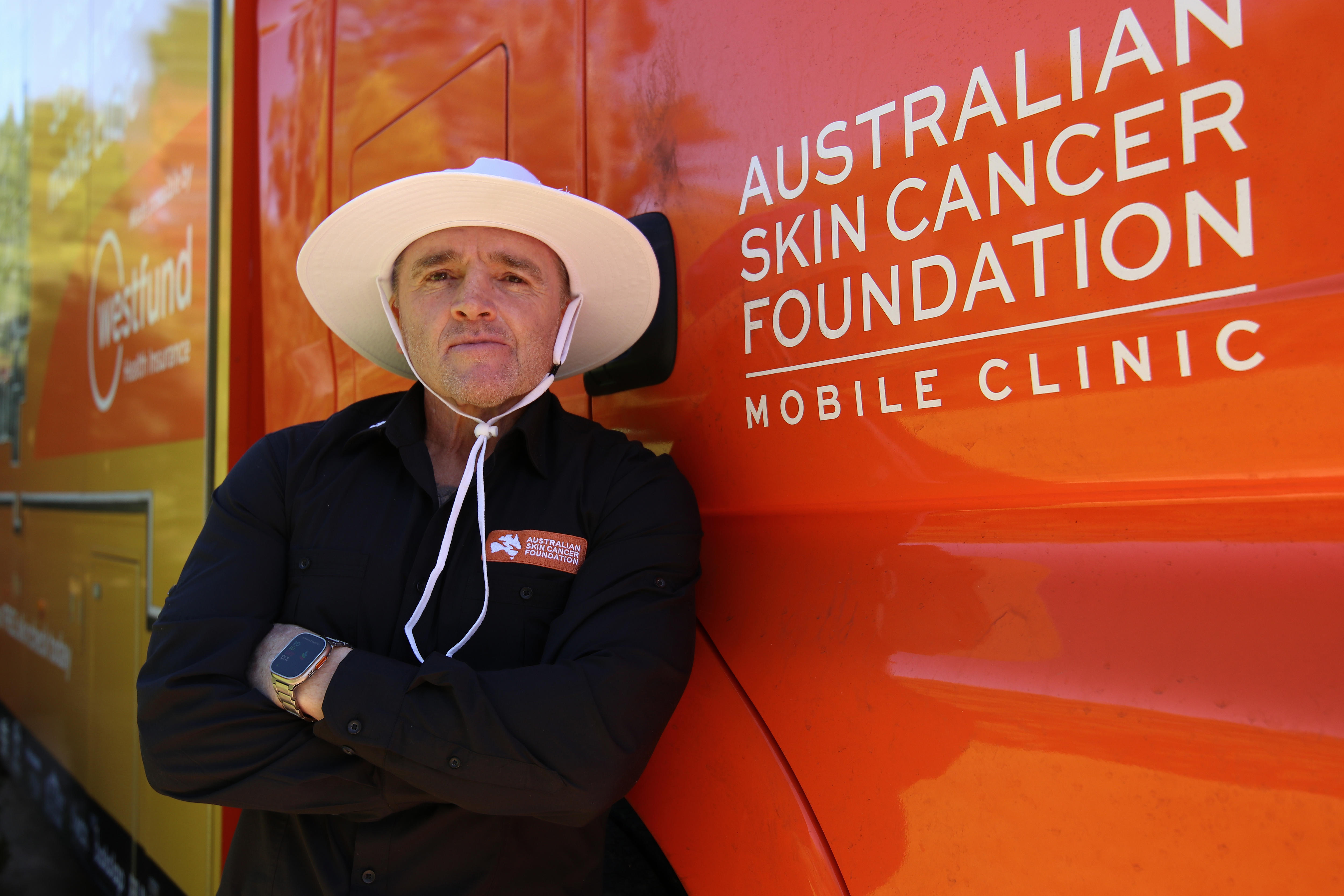Jay Allan wearing a long sleeve black shirt with a wide brim hat leaning against orange and yellow van with arms crossed.