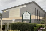 The outside of a one story grey building with a sign and grass outside on a grey day.