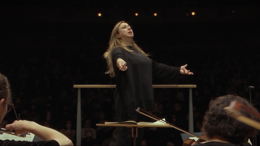 White middle-aged woman with long light brown hair is dressed in all black and conducts an orchestra from a conductor's podium