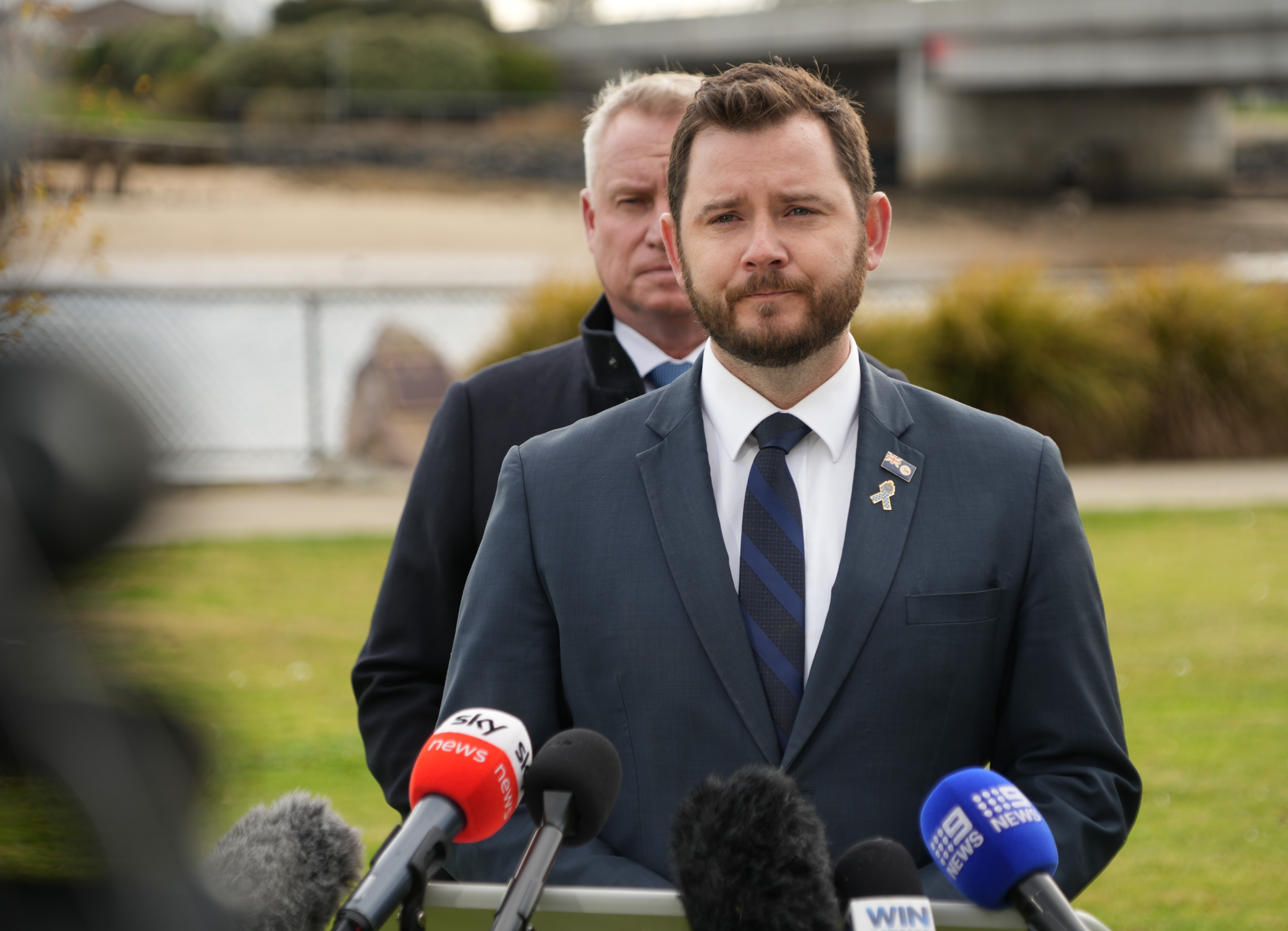 Two men in suits stand on a football field in front of microphones, addressing the media.