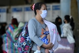 A woman wearing a face mask carries a baby at a bus station.