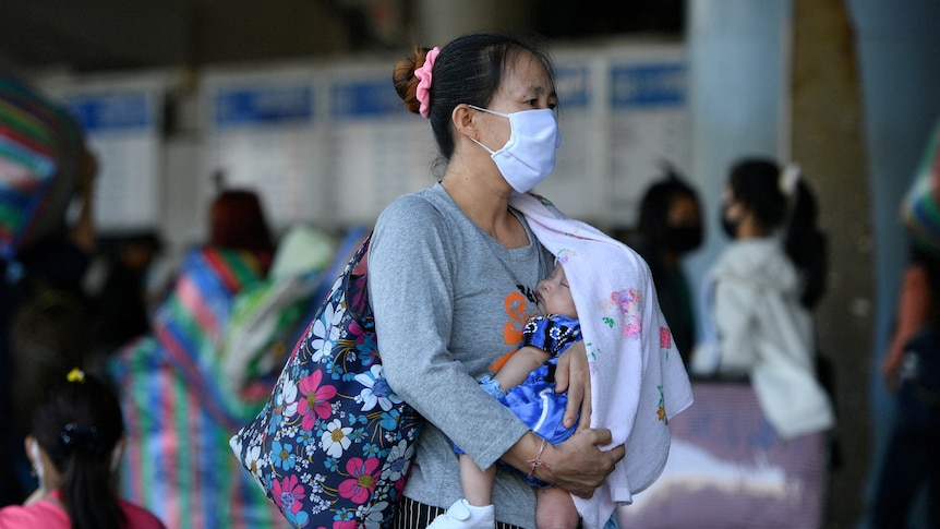A woman wearing a face mask carries a baby at a bus station.