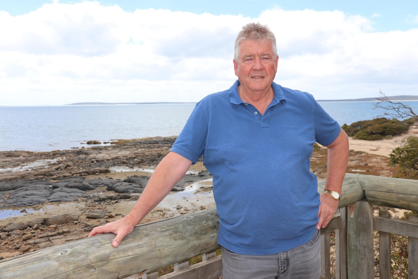 A middle-aged man leans on a wooden railing in front of a rocky beach.