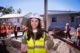 A smiling woman with long, dark hair holds a shovel across her shoulder at a construction site. She wears a hard hat.