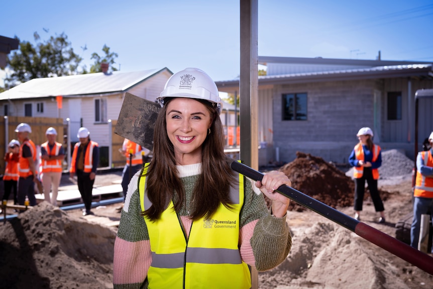 Una mujer sonriente con cabello largo y oscuro sostiene una pala sobre su hombro en un sitio de construcción.  Ella usa un casco.