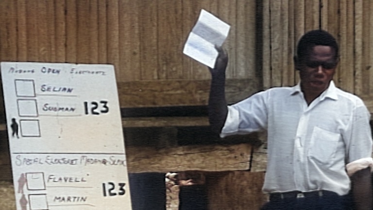 Outside, in front of a stilted wooden building, a man demonstrates how to vote using a large replica of the ballot paper.