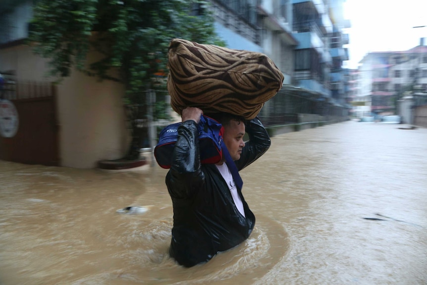 A man wades through yellow-brown floodwaters on a street lined with medium-density apartment buildings as he carries two bags.
