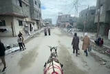A donkey wearing a red harness walks behind several people with piles of rubble on either side of it.