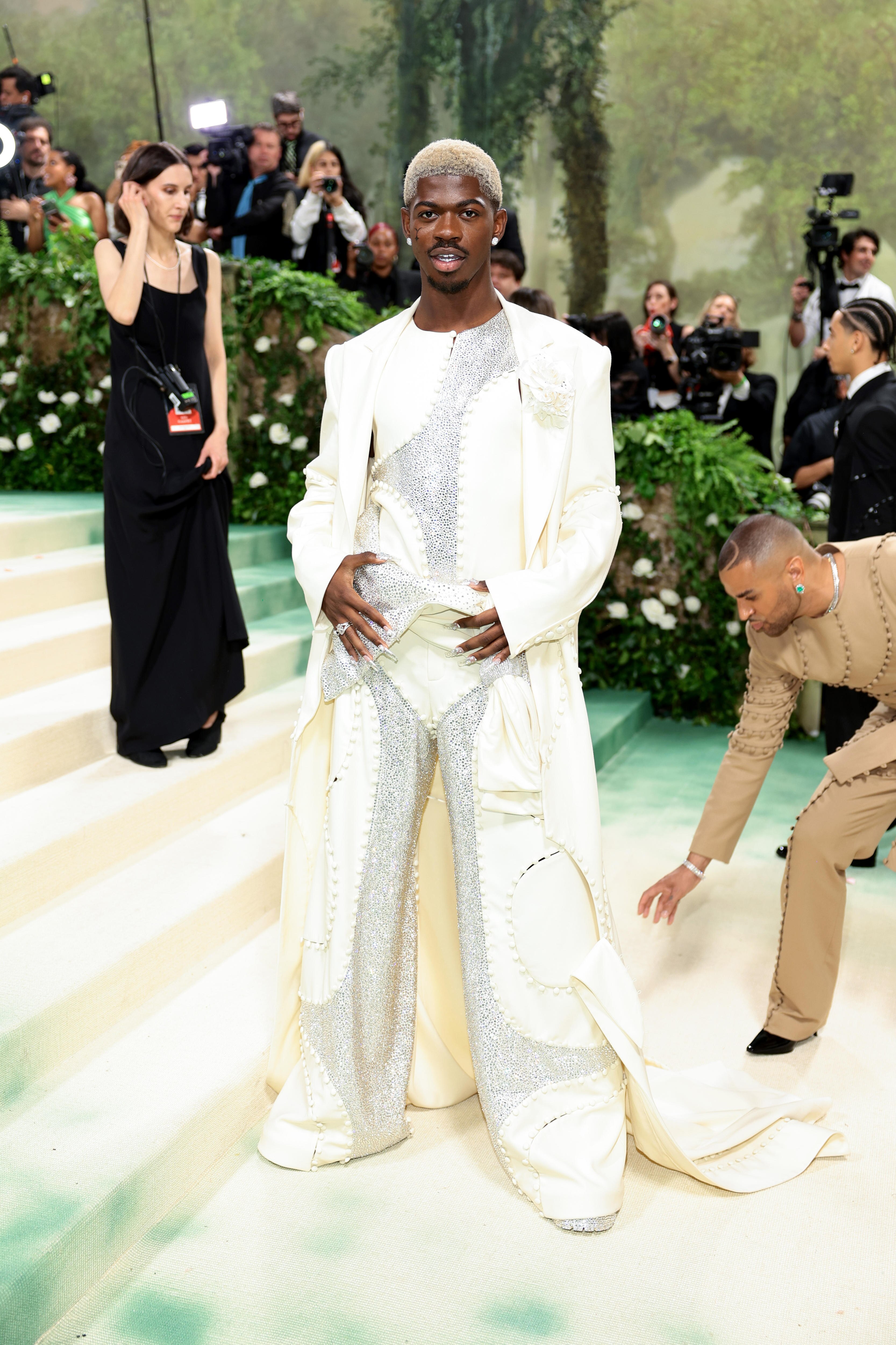 Lil Nas X stands in an all-white outfit of pants, coat and shirt, adorned with diamonds. His hair is buzzcut and bleached.