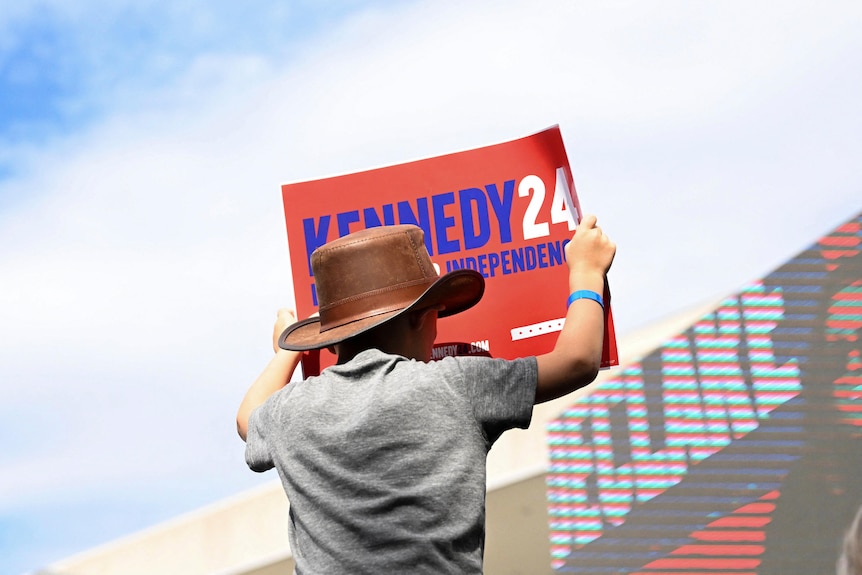 A person in a hat holds up a sign reading "Kennedy 24"