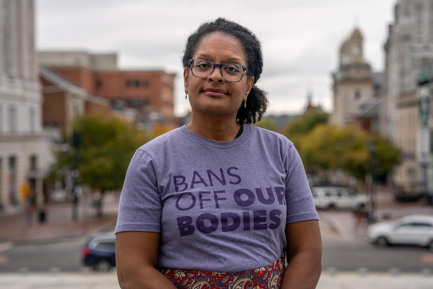 A black woman in glass stand in the street in a purple T-shirt that reads "bans off our bodies"
