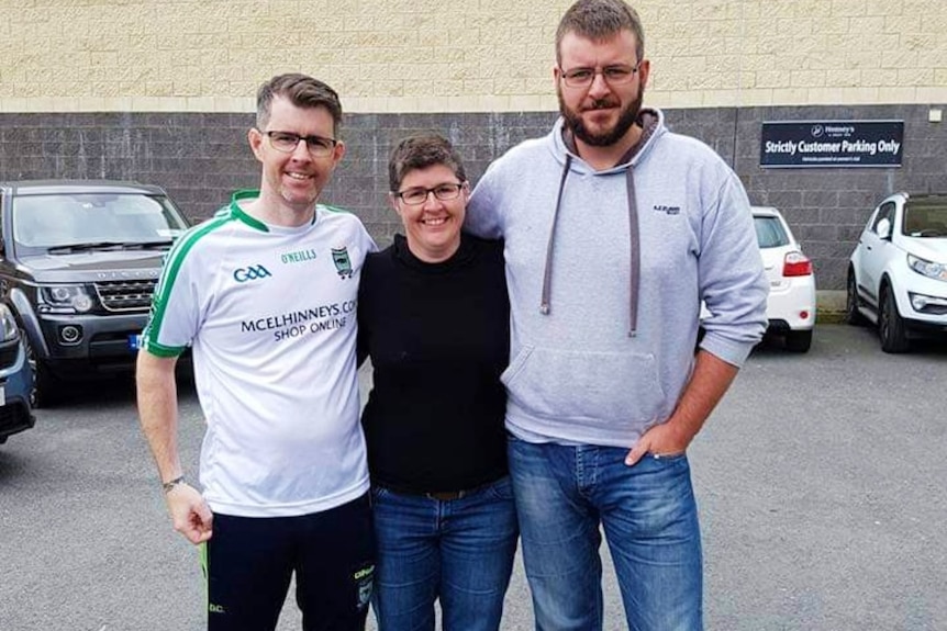 A woman stands arm in arm with two men in a car park
