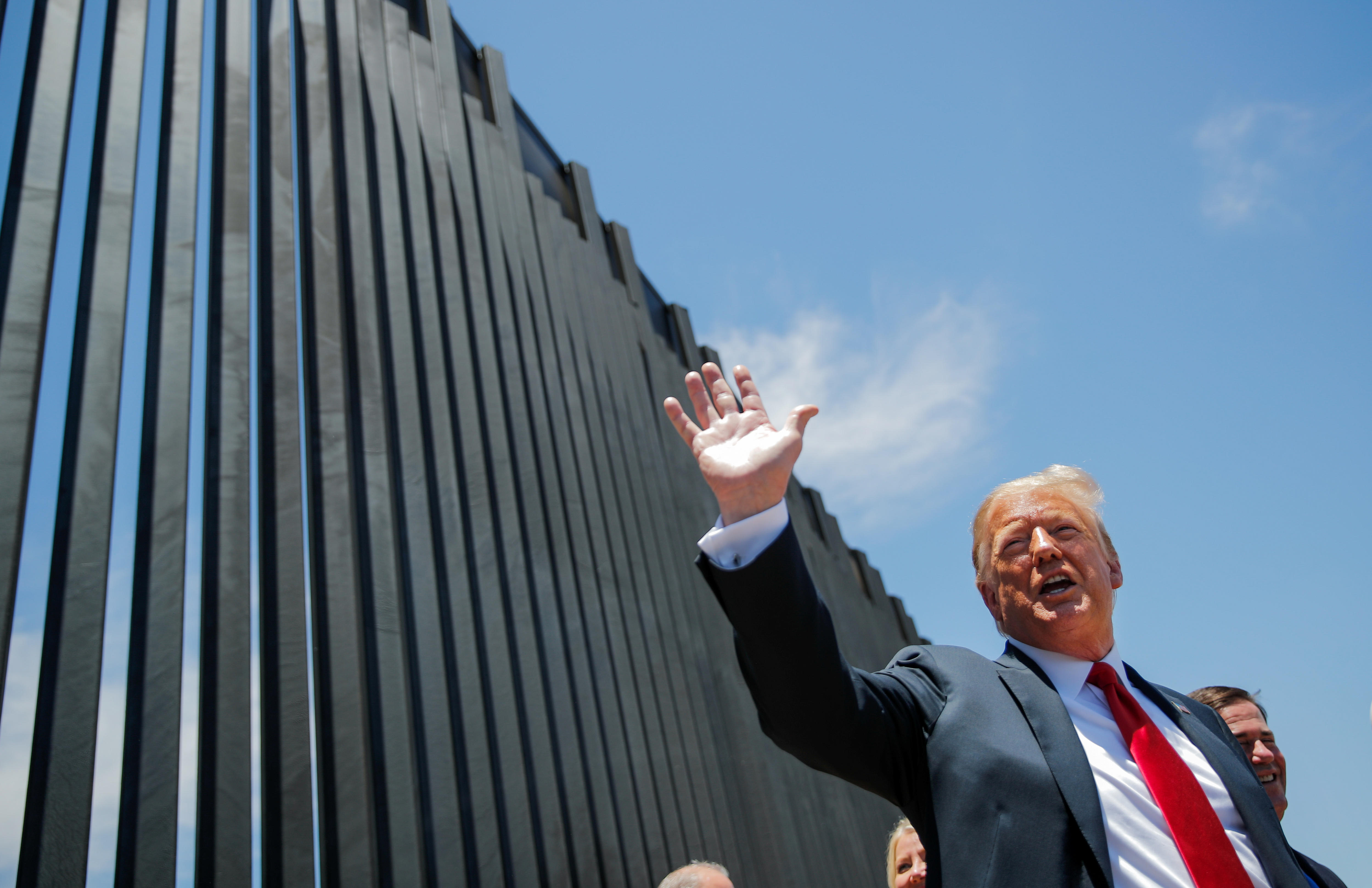 Donald Trump wears a black suit and red tie and stands by the thick bars of a border wall, about to sign a plaque.