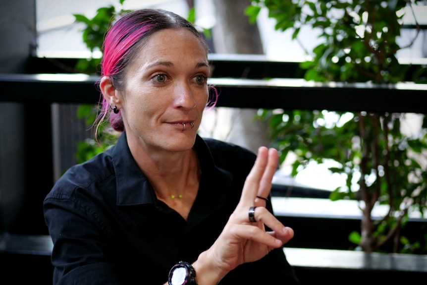 A close up of a woman with pink hair and lip piercings, she is wearing a black shirt and making a hand sign with two fingers up