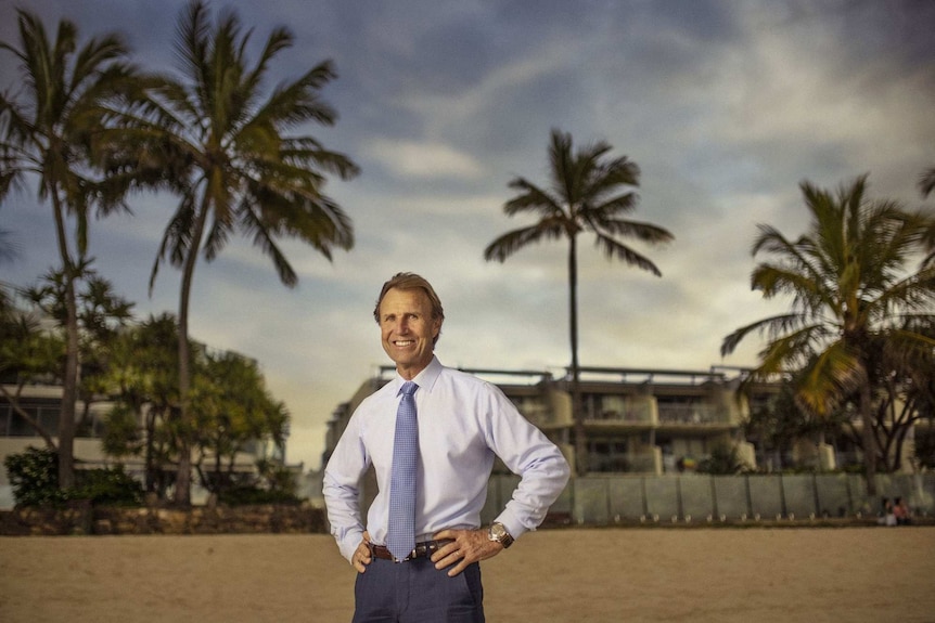 A man in a suit and tie stands on a beach with his hands on his hips and a to storey beachfront house behind himself.
