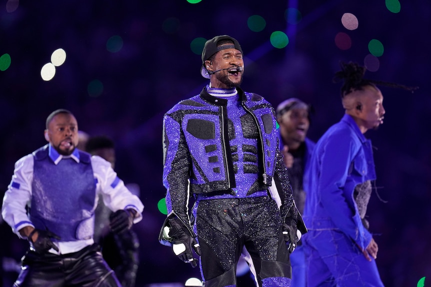 Usher wearing a sparkly black and blue armoured jacket, singing on stage