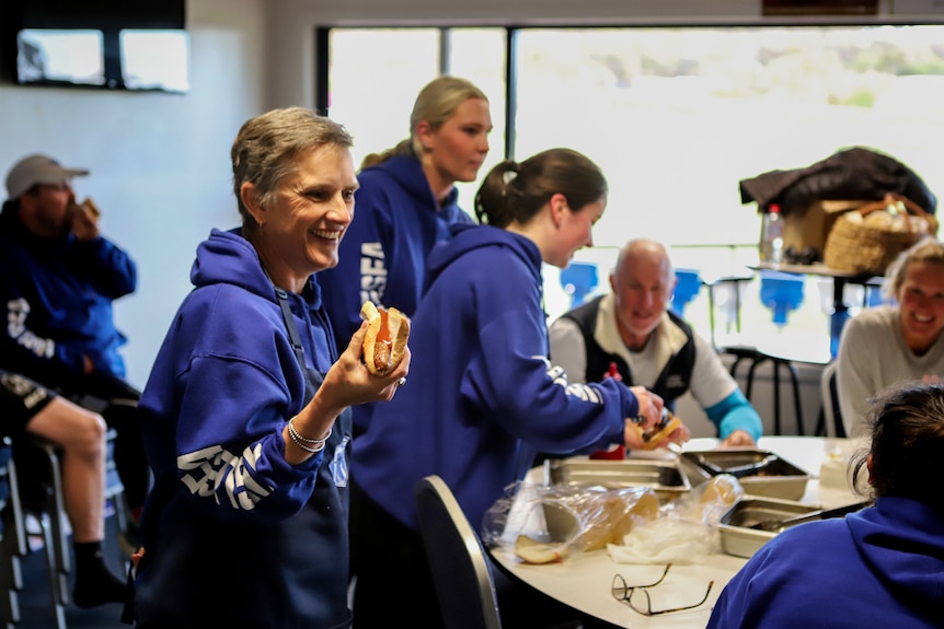 Women in blue hoodie holds a sausage in bread with other people eating sausages in a room behind her