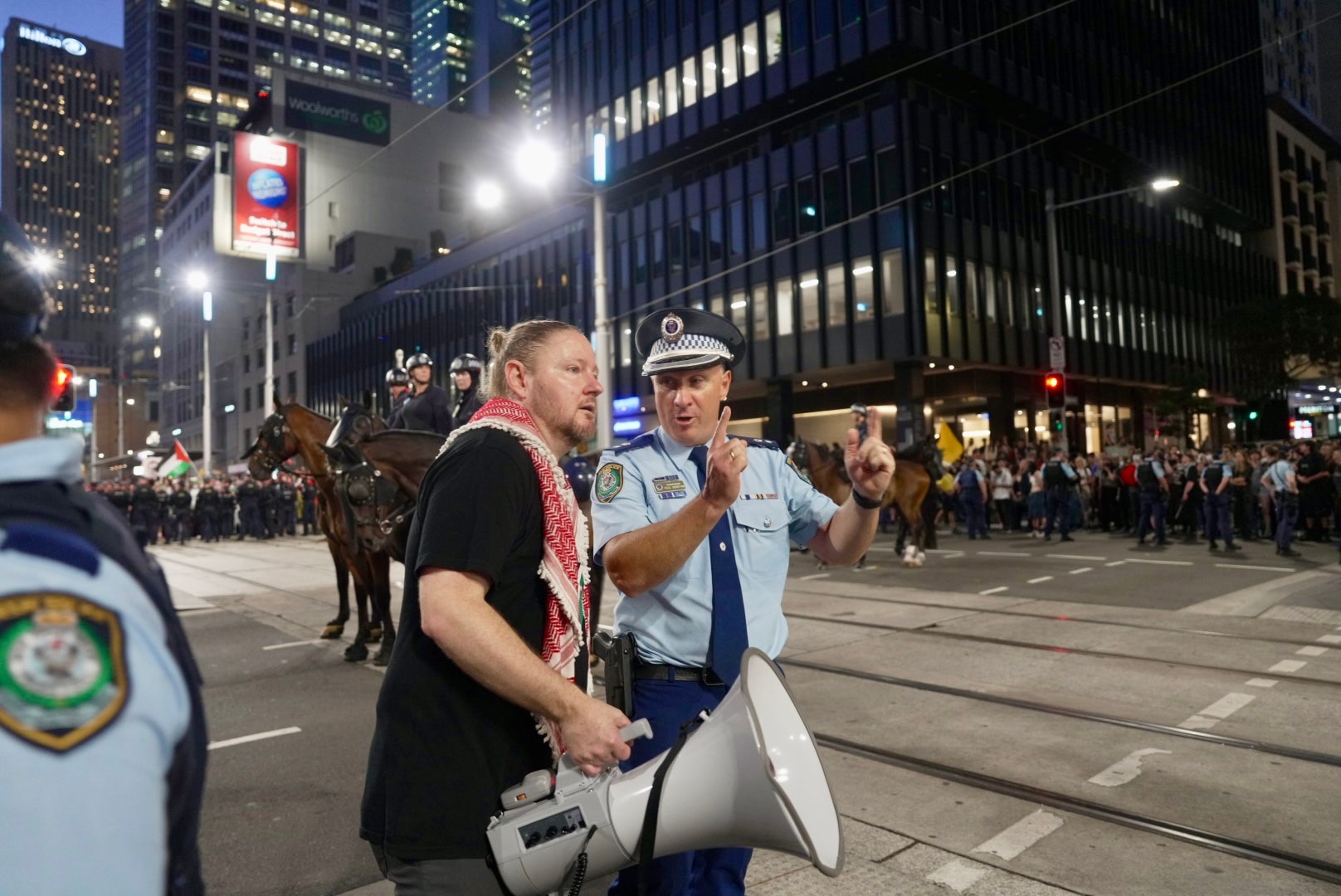 Hombre con camisa negra y un gran megáfono hablando con un oficial de policía en una calle oscura y concurrida de la ciudad
