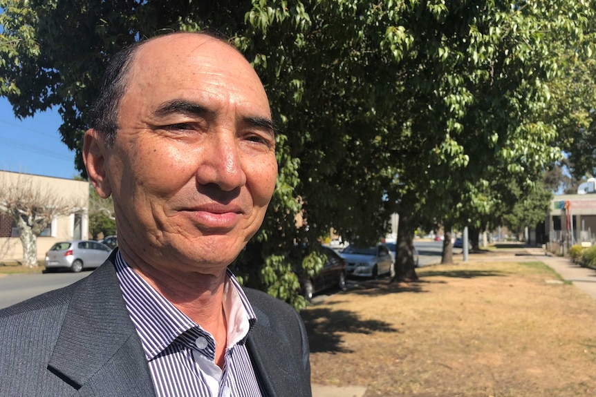 A man in a suit smiles at the camera with trees in the background.