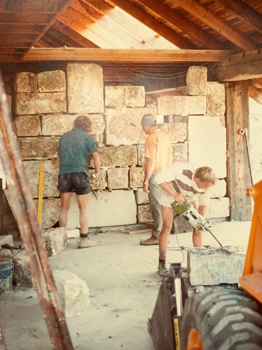 Three men work on building a stone block wall in a house at Logan.