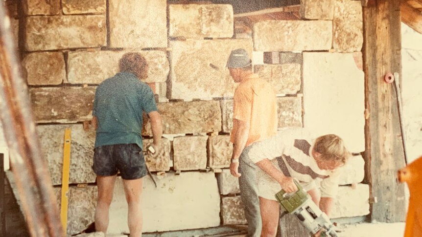 Three men work on building a stone block wall in a house at Logan.