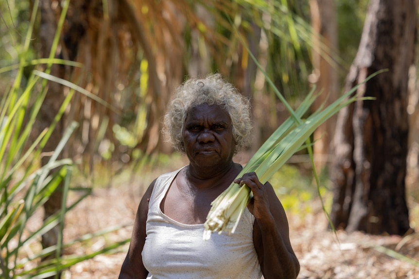 Doreen Jinggarrabarra, a Burarra woman with dark brown skin and grey curly hair holds pandanus leaves in the bush.