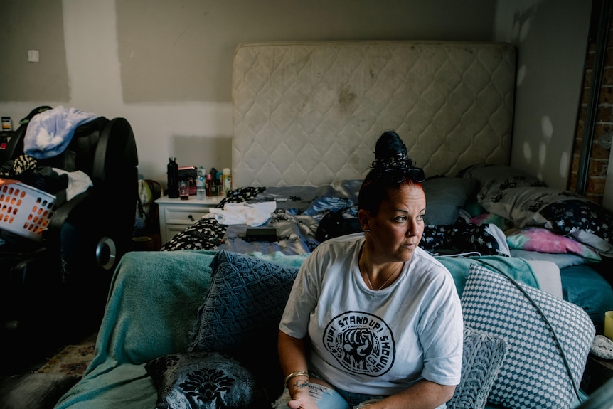 A woman sits on a couch in a garage filled with furniture and clothes. Behind her a mattress is against a wall.