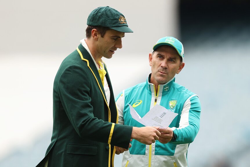 A man in a green jacket and hat is handed a paper list from another man in a light green tracksuit