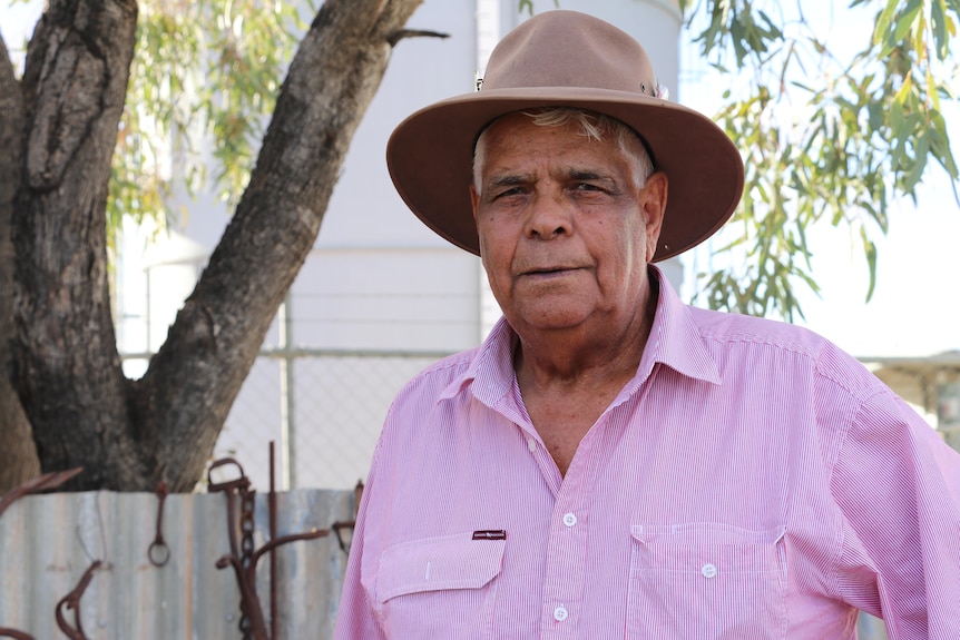 An Aboriginal elder stands outside in a pink shirt and beige hat.