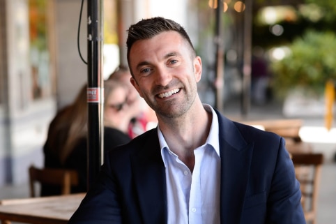 Adelaide city councillor Robert Simms sits at a cafe's outdoor tables with a coffee, smiling at the camera.