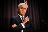 Malcolm Turnbull speaks at a lectern in front of a blue curtain. He is frowning and holding his glasses in one hand.