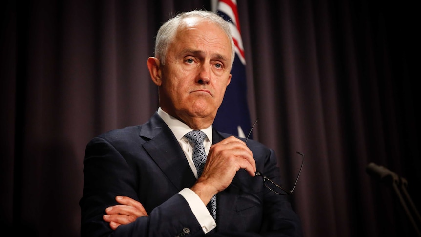 Malcolm Turnbull speaks at a lectern in front of a blue curtain. He is frowning and holding his glasses in one hand.