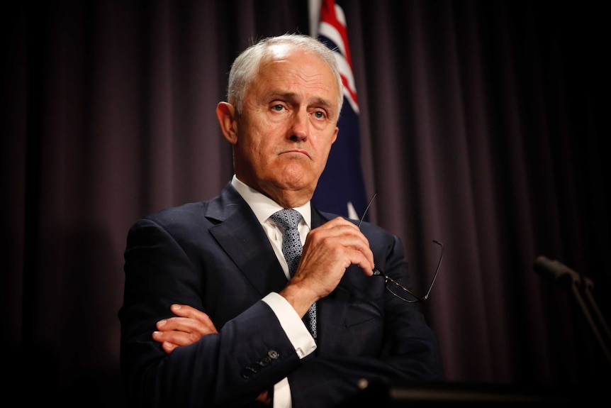 Malcolm Turnbull speaks at a lectern in front of a blue curtain. He is frowning and holding his glasses in one hand.