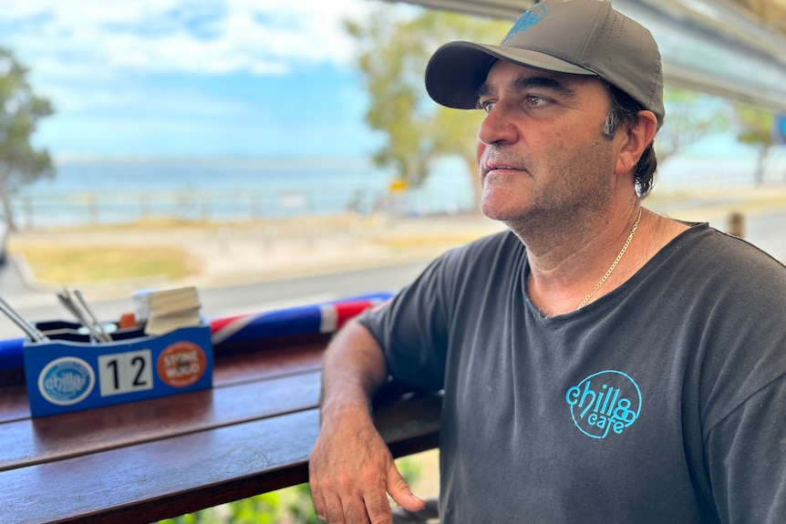 A man leaning on a bench at a beachside cafe. He is wearing a baseball cap and t-shirt.