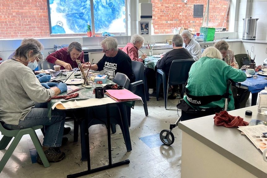 A group of older, female artists gather around indoor tables spread with art supplies, the sun shining in from a big window