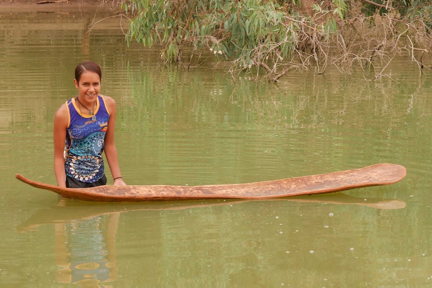 Mununjali/Wiradjuri girl Shanaha Clayton stands in the Darling River in Wilcannia, beside an Aboriginal canoe