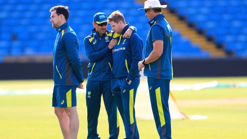 Justin Langer puts his arms around Steve Smith, who is looking down with his eyes closed, both wearing blue training kit
