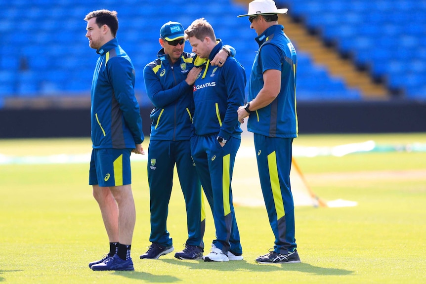 Justin Langer puts his arms around Steve Smith, who is looking down with his eyes closed, both wearing blue training kit