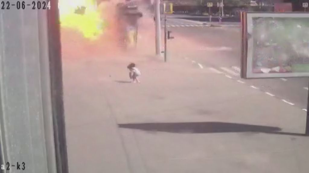 A woman holding a shopping bag walks slowly along a wide footpath before a blast and fireball erupts near her