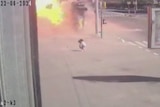 A woman holding a shopping bag walks slowly along a wide footpath before a blast and fireball erupts near her