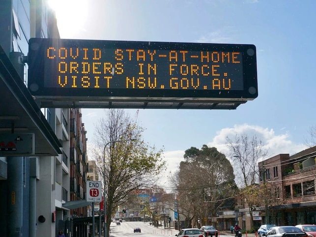 A large sign advises stay at home orders are in force in the foreground of an empty, sunny street