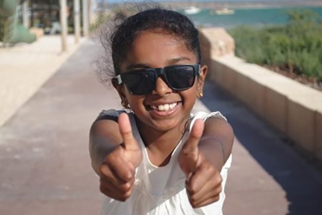 Head and shoulders of a young girl smiling, wearing sunglasses and holding two thumbs up