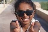 A head and shoulders of a young girl smiling, wearing sunglasses and holding two thumbs up