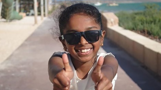 A head and shoulders of a young girl smiling, wearing sunglasses and holding two thumbs up