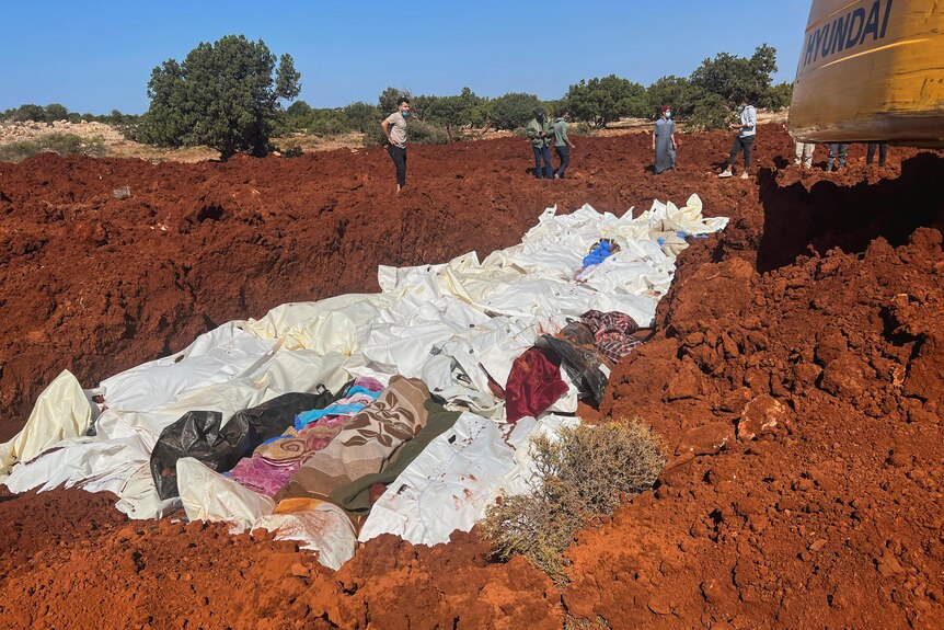 Bodies wrapped in blankets are placed into a mass grave, an excavator can be seen in upper right corner