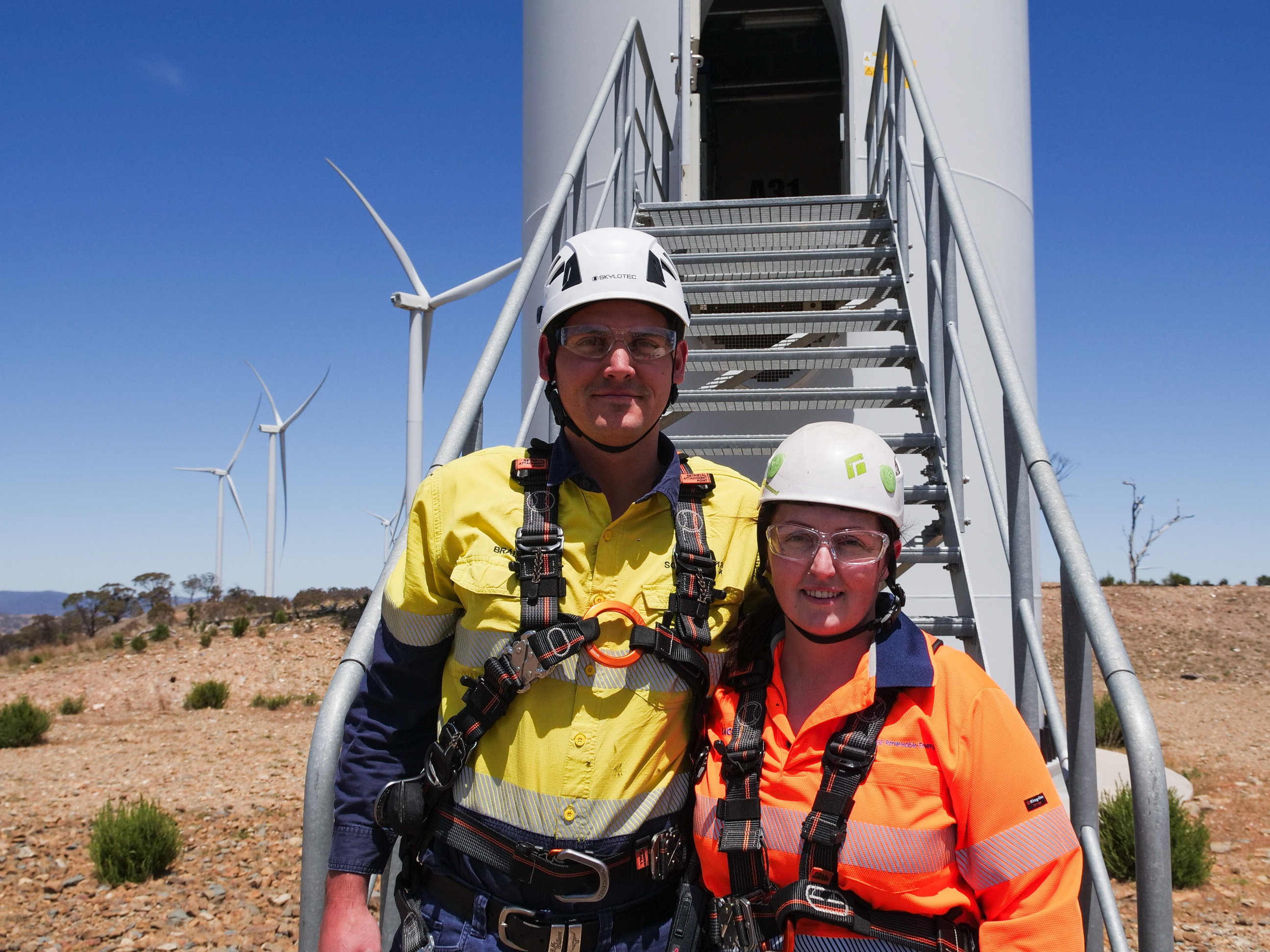 A man and a woman in high vis and helmets stand together at the base of a wind turbine.