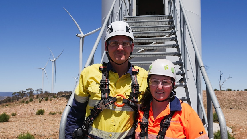 A man and a woman in high vis and helmets stand together at the base of a wind turbine.