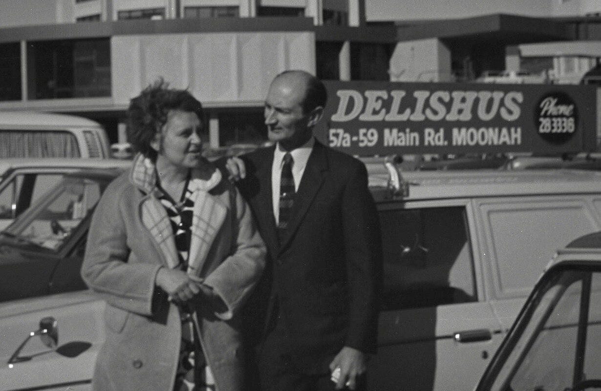 Black and white image of man and woman standing in front of advertisement on car reads "Delishus"